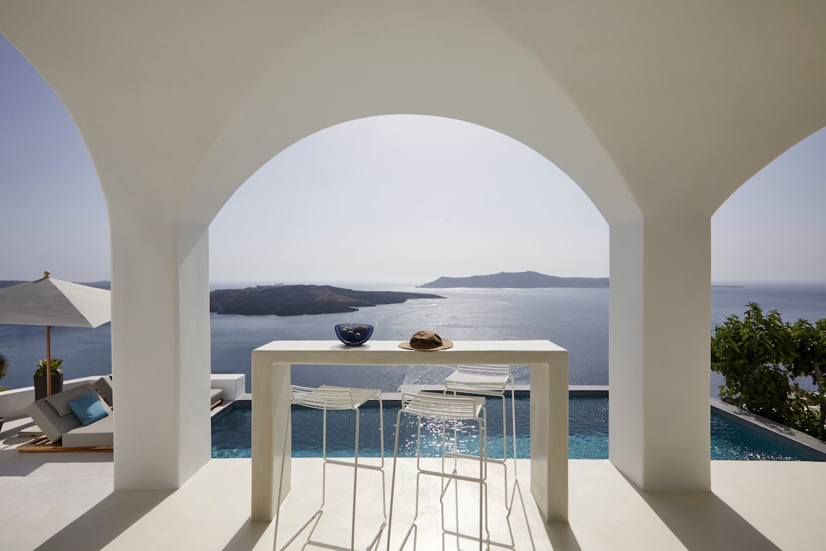 An outdoor terrace features a sleek, modern table with three white bar stools, overlooking the serene Aegean Sea. An umbrella provides shade to a nearby lounge area, while the architecture showcases elegant arches framing the scenic caldera views.