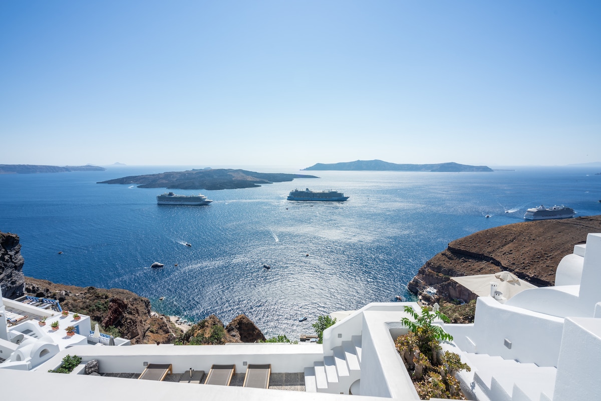 A panoramic view showcases the deep blue Aegean Sea, dotted with boats, framed by the rugged coastline. The scene highlights the vibrant colors of the water against the clear sky, with islands visible in the distance, enhancing the sense of tranquility and connection to nature.