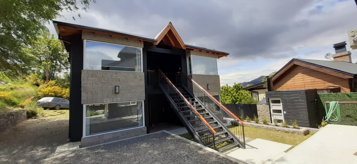 A two-story building features a modern architectural design, with large windows allowing natural light to fill the space. A staircase with a contrasting orange railing leads to the entrance. The exterior is complemented by a gravel driveway and landscaped areas.