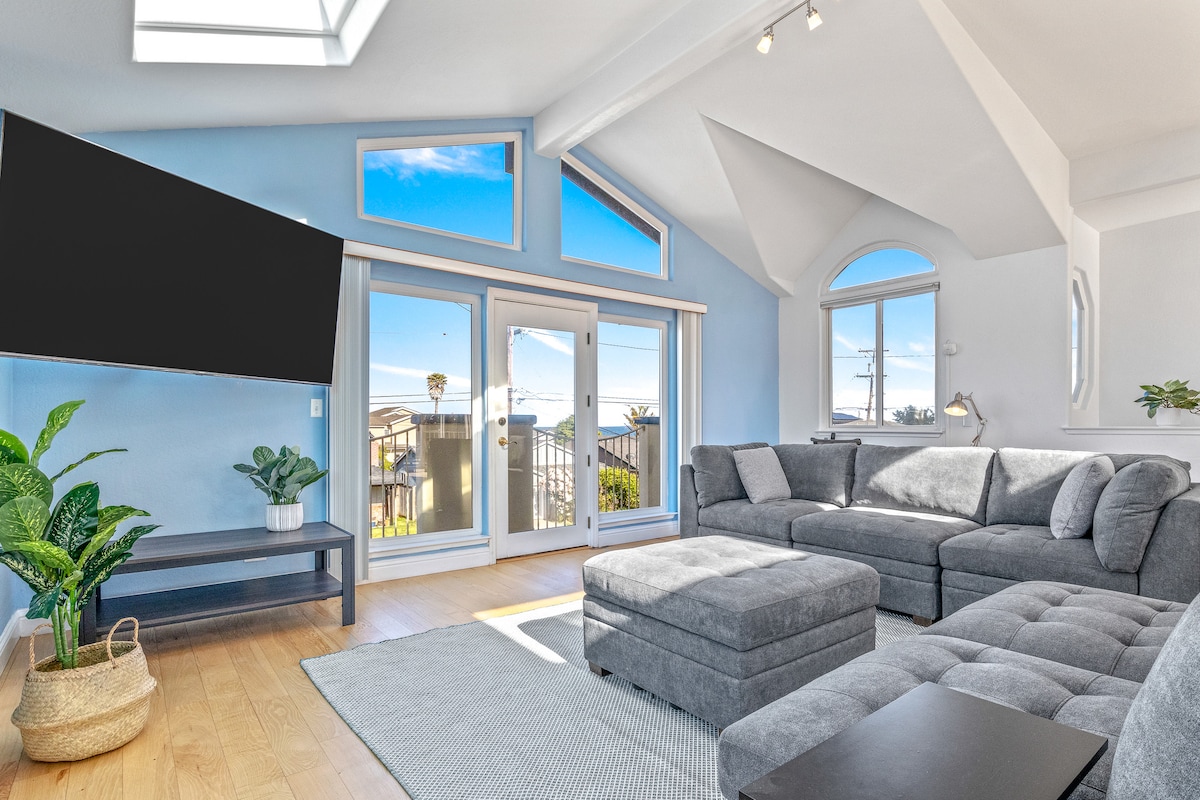 A spacious living area is illuminated by natural light streaming through large windows. A comfortable sectional sofa and an ottoman are positioned on a light-colored rug, while a modern television is mounted on the wall. Potted greenery enhances the inviting atmosphere.