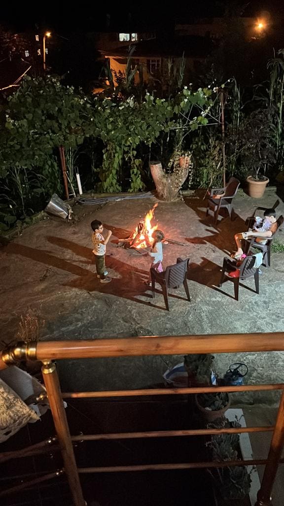 A gathering is taking place around a small campfire, with five individuals seated on chairs arranged in a circular formation. The warm glow of the fire contrasts against the darker surroundings, illuminated by greenery and night lights in the background.