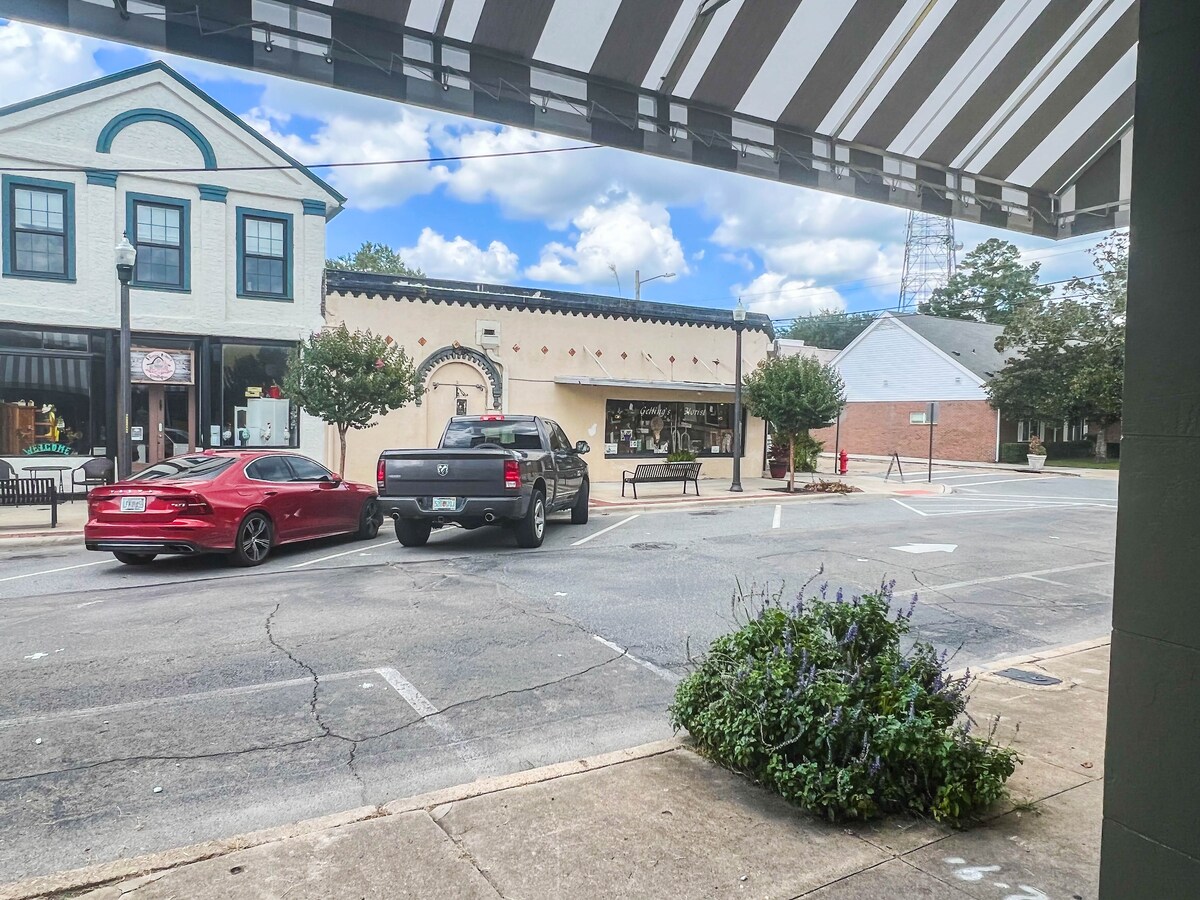 A view of the vibrant downtown street reveals a mix of shops and eateries across the way. Two parked vehicles are visible, alongside a planter filled with greenery. Striped awnings provide shade, and a clear blue sky with scattered clouds adds to the scene.