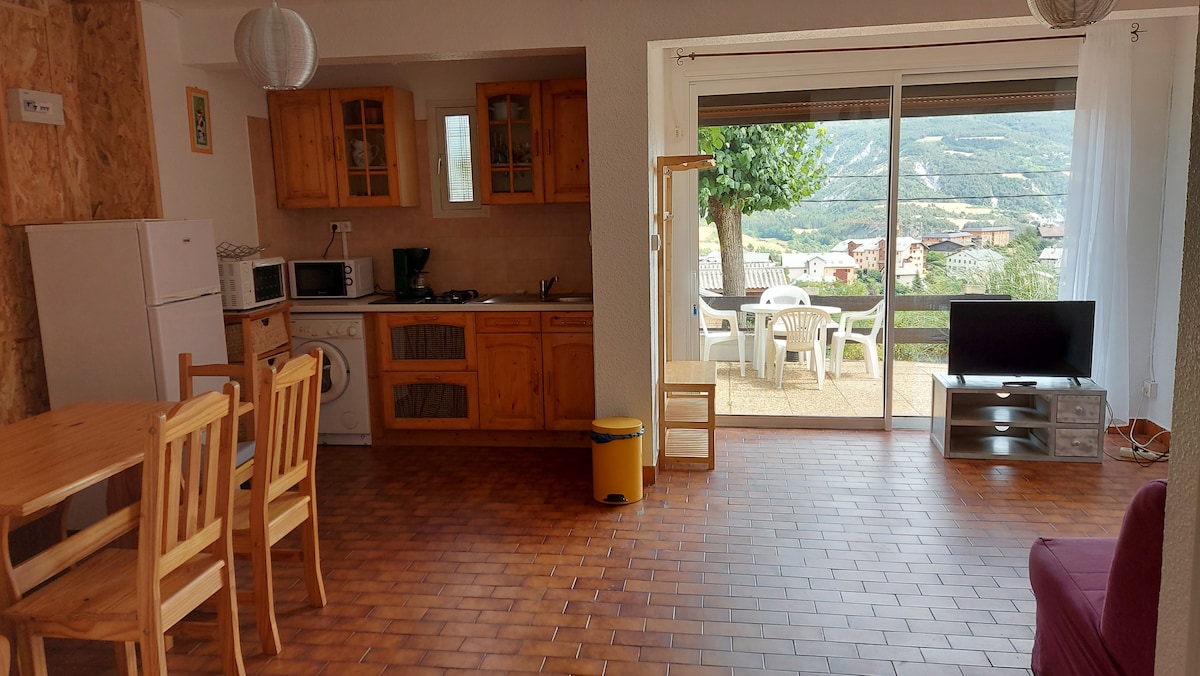 An open living space features wooden cabinetry, a dining table with four chairs, and a kitchenette equipped with essential appliances. Large glass doors provide access to a private terrace, offering views of the surrounding landscape. A comfortable seating area includes a television and colorful sofa.