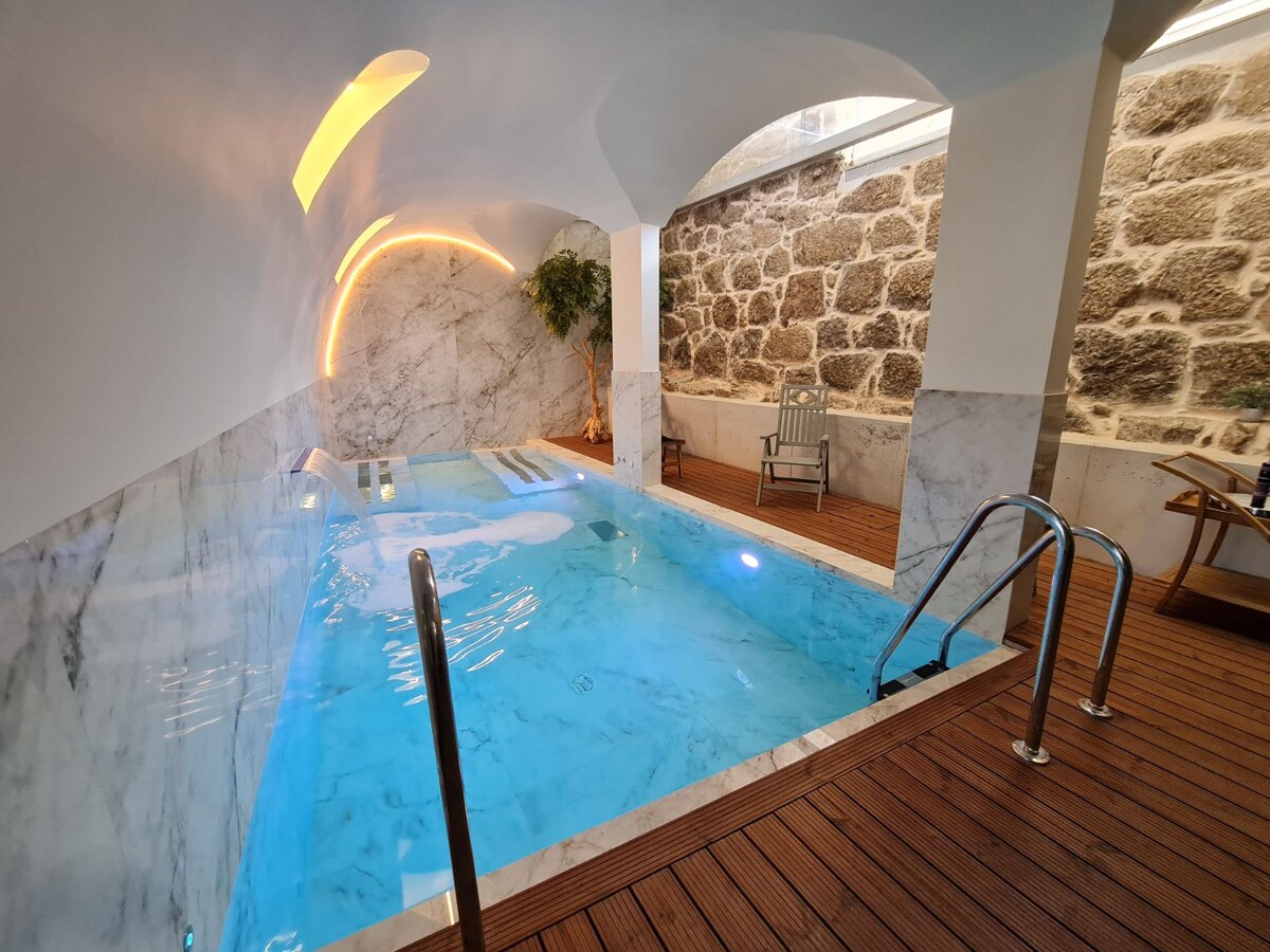 An indoor spa pool is set against a backdrop of textured stone walls. Soft lighting accentuates the water, creating a calming ambiance. A wooden deck surrounds the pool, and a comfortable seating area is visible, offering a place for relaxation.