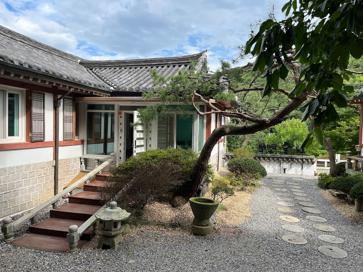 A serene pathway meanders through a garden featuring meticulously arranged stones and greenery. Traditional architecture is highlighted by a slate roof and wooden accents. A decorative stone lantern is present, while a mature tree adds natural character to the inviting exterior.
