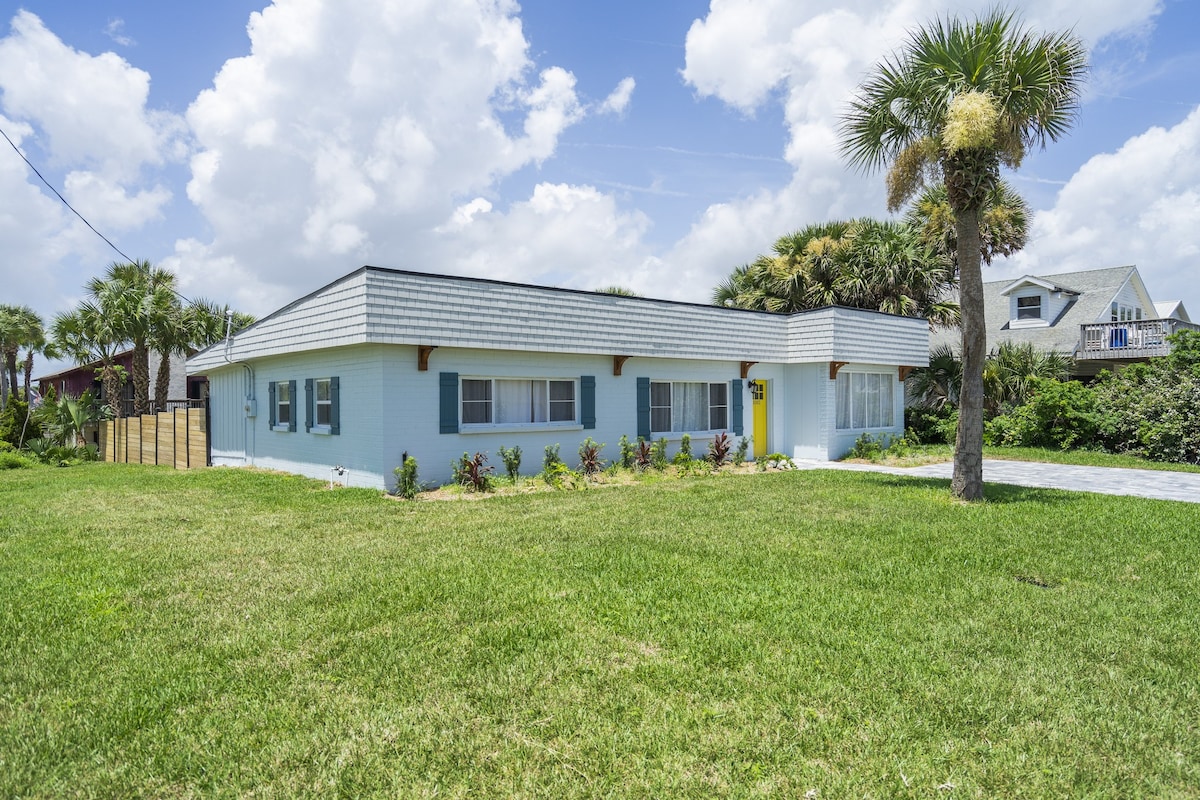 A single-story blue house is framed by lush green grass and palm trees. The front features large windows and a vibrant yellow door, complemented by well-maintained landscaping. A clear sky with scattered clouds adds to the inviting atmosphere.