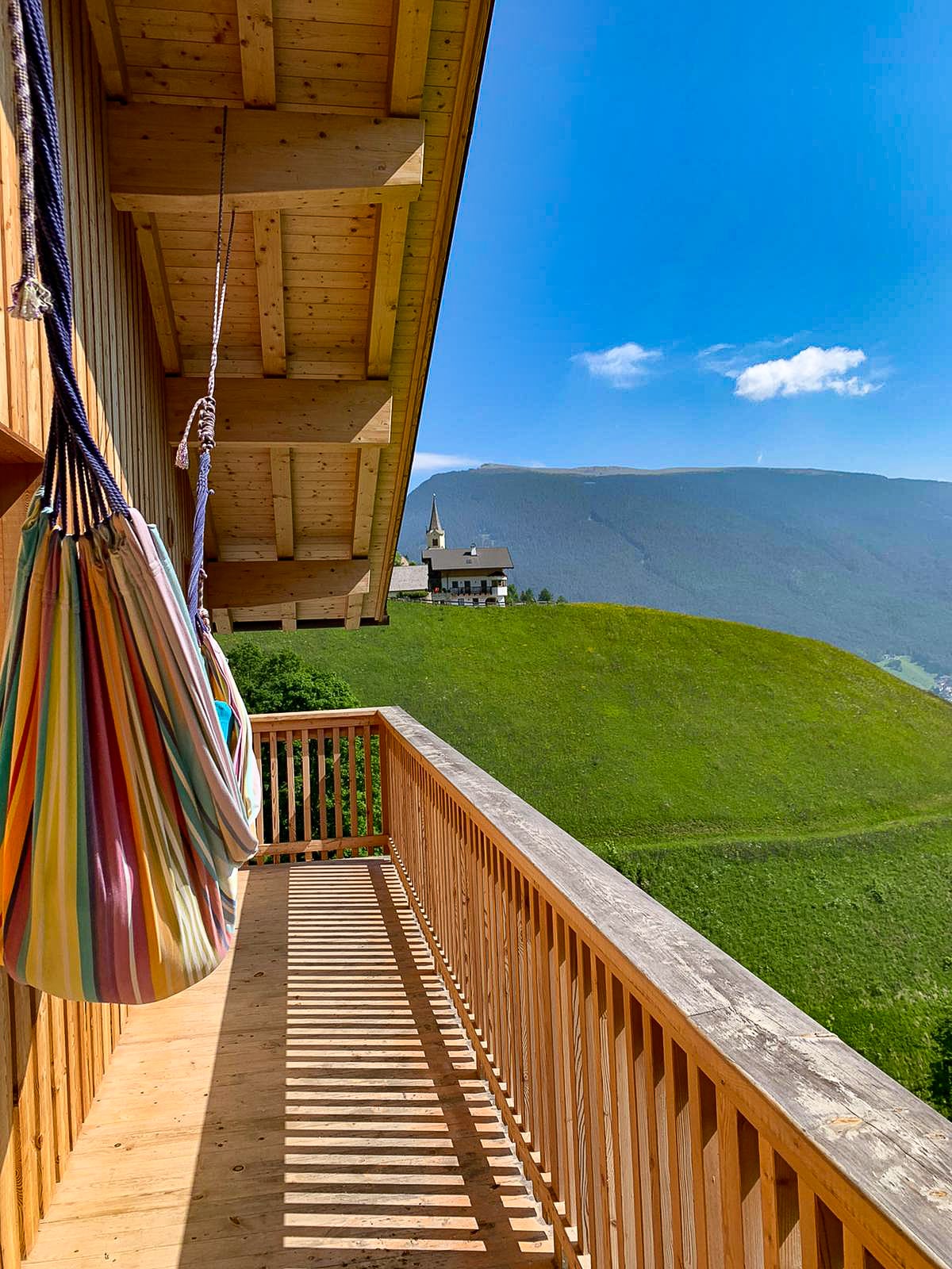 A wooden balcony features colorful hammocks suspended from the ceiling. A sloping green hill is visible in the background, leading to a distant church steeple. The sky is clear with a few clouds, emphasizing the tranquil mountainous landscape.