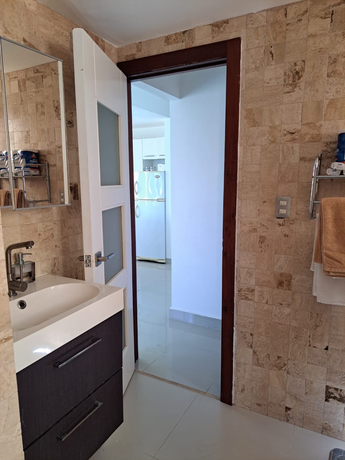 A small bathroom is shown with a modern sink and cabinetry in dark hues. Light-colored stone walls add texture, while a mirror is mounted above the sink. A door leads to another room, partially visible, indicating an open layout.