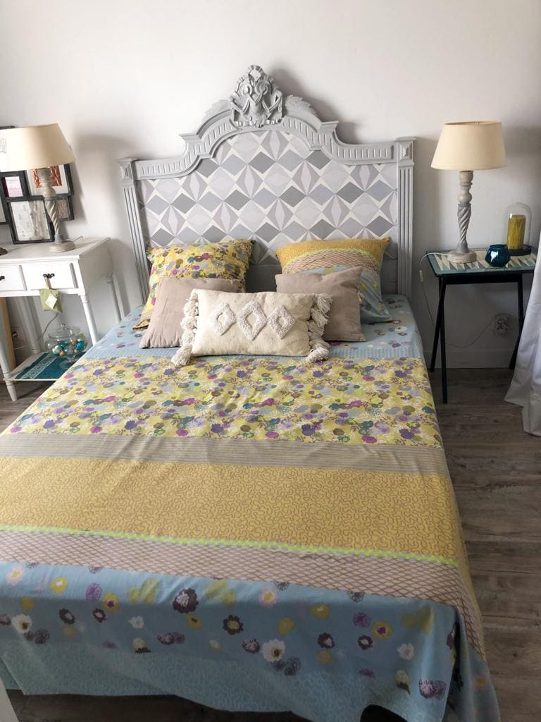 A cozy bedroom is presented with a patterned bedspread featuring vibrant floral designs. Decorative pillows are arranged on the bed, which is framed by a stylish headboard. A bedside lamp and a small table complement the space, enhancing its welcoming feel.