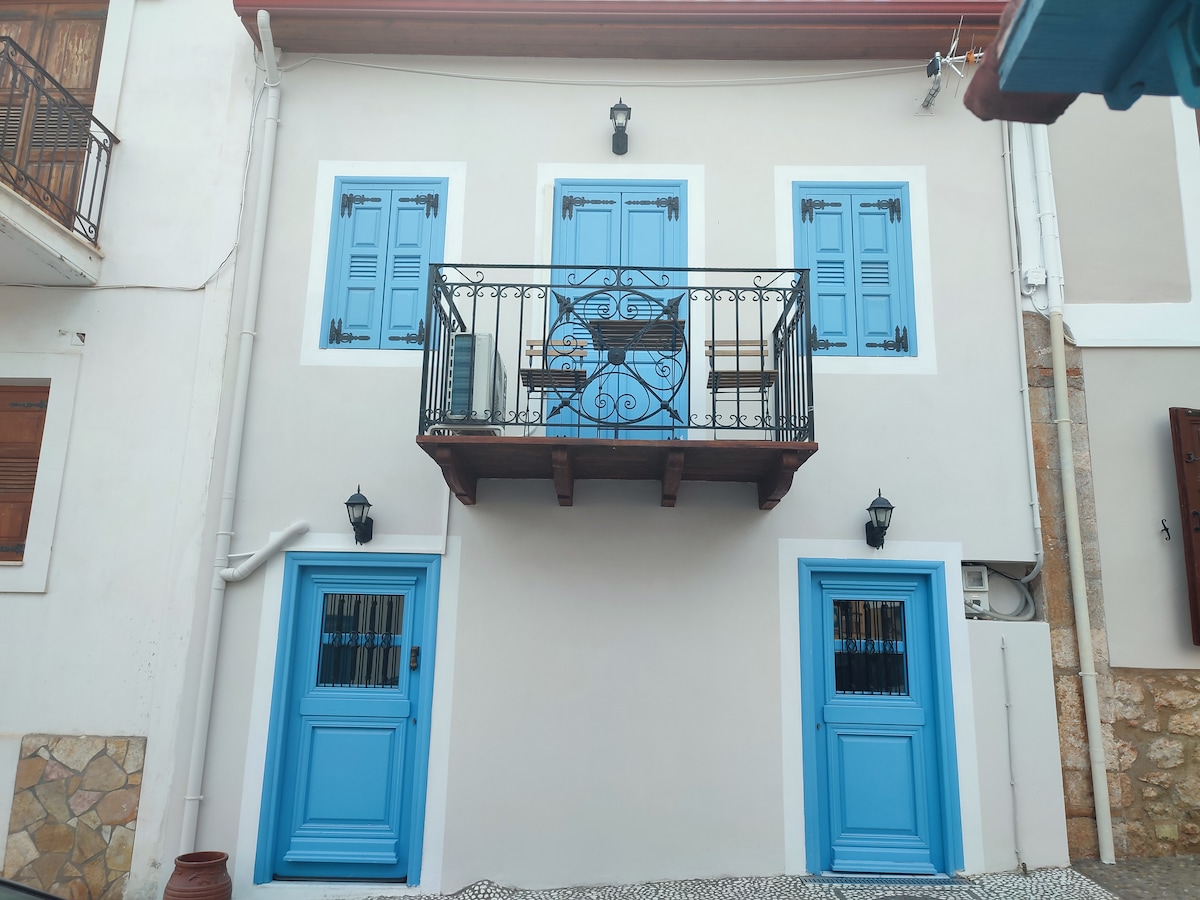 The exterior of a charming building features light-colored walls and blue shutters. A balcony with decorative ironwork is situated above the entrance, which is flanked by two matching doors. Wall sconces provide soft lighting, enhancing the welcoming look of the façade.