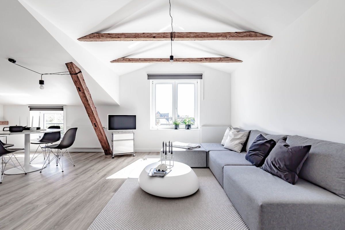 A bright and modern living area features exposed wooden beams and large windows allowing natural light to fill the space. A cozy gray sectional sofa is paired with a round coffee table, while a small dining area is visible in the background, complete with sleek chairs.
