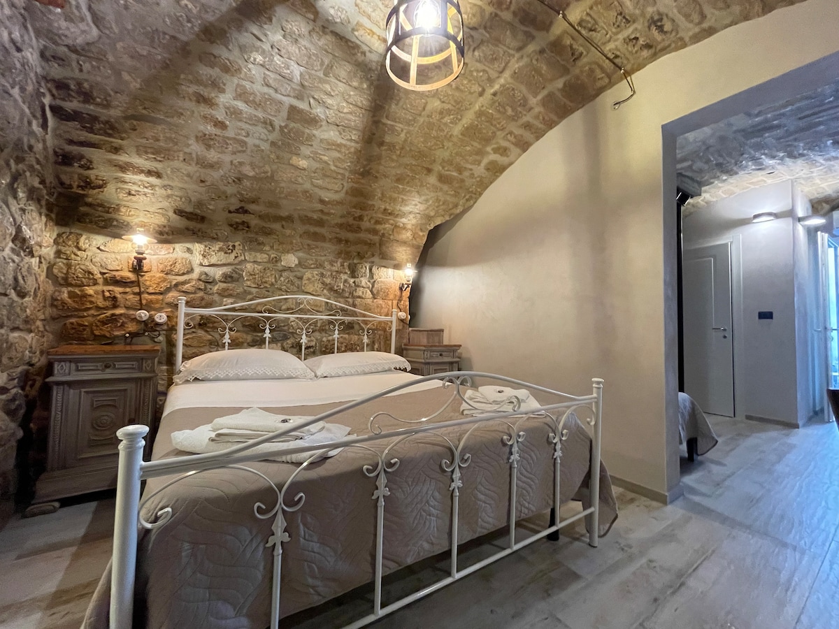A spacious bedroom features a stone arched ceiling and soft lighting. A white wrought-iron bed is dressed with neutral-toned linens. Wooden nightstands flank the bed, adding a rustic touch. The warm hues of the stone walls create a calming ambiance.