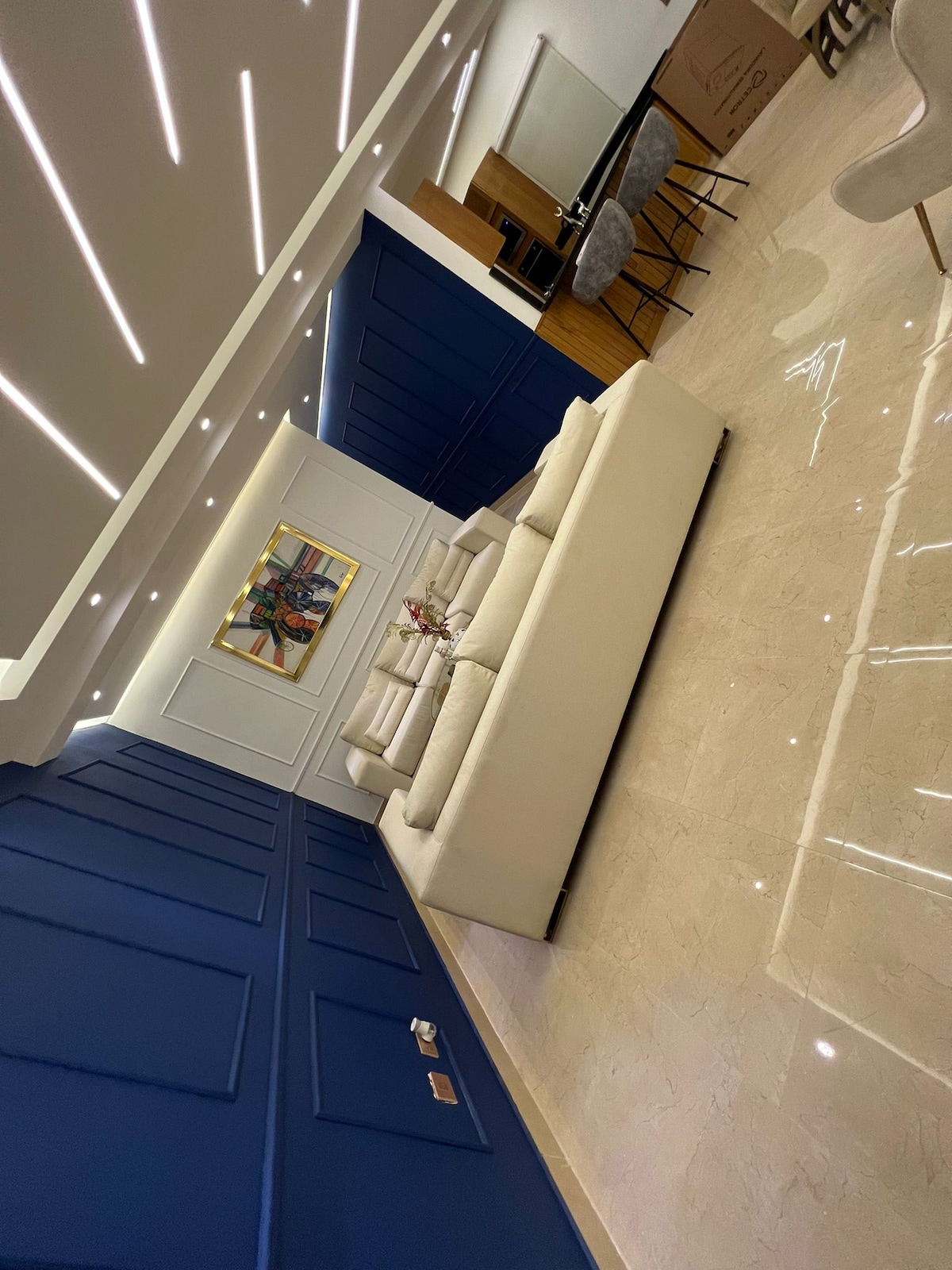 A spacious living area features a light-colored sectional sofa centered against a deep blue accent wall. Elegant artwork hangs above, complemented by modern recessed lighting along the ceiling. A polished marble floor enhances the room's contemporary aesthetic.
