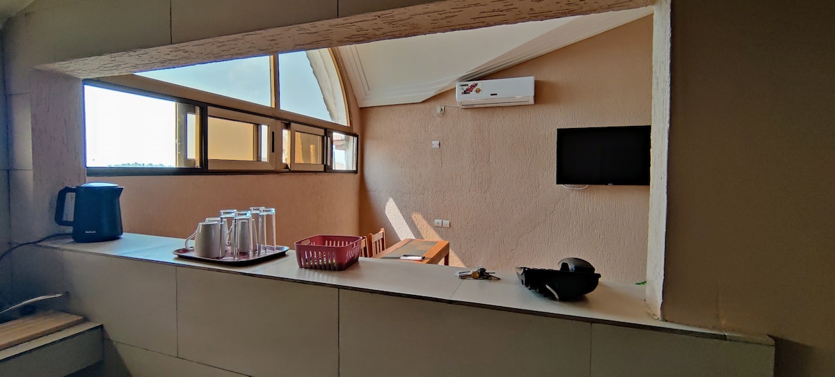 A kitchen space is visible through an open countertop. The scene includes a kettle, a tray with multiple glasses, a basket, and a plate. Beyond, a dining area is seen with a wooden table and chairs, accompanied by a wall-mounted television and a large window allowing natural light.