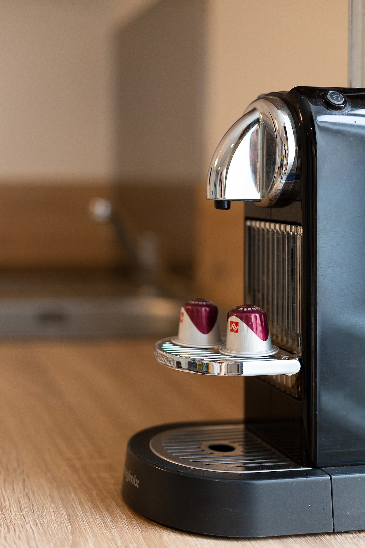 A sleek coffee machine sits on a wooden counter, displaying two coffee capsules ready for use. The background features soft, neutral tones, providing a subtle, contemporary kitchen space.