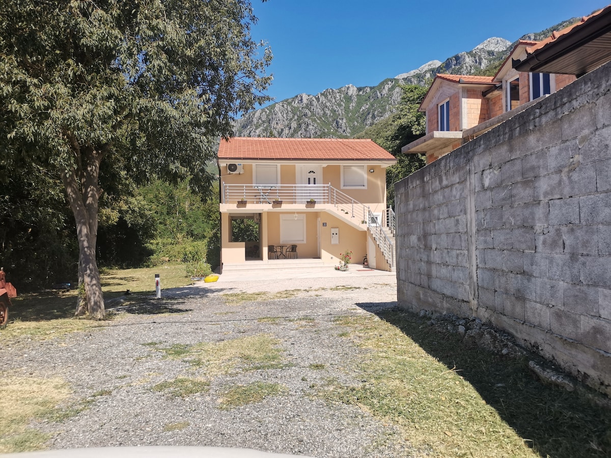 The two-story apartment building is positioned against a backdrop of mountains, showcasing a spacious outdoor area with gravel parking. A staircase leads to an upper terrace, while trees provide shade nearby. The facade features large windows and a welcoming entrance.