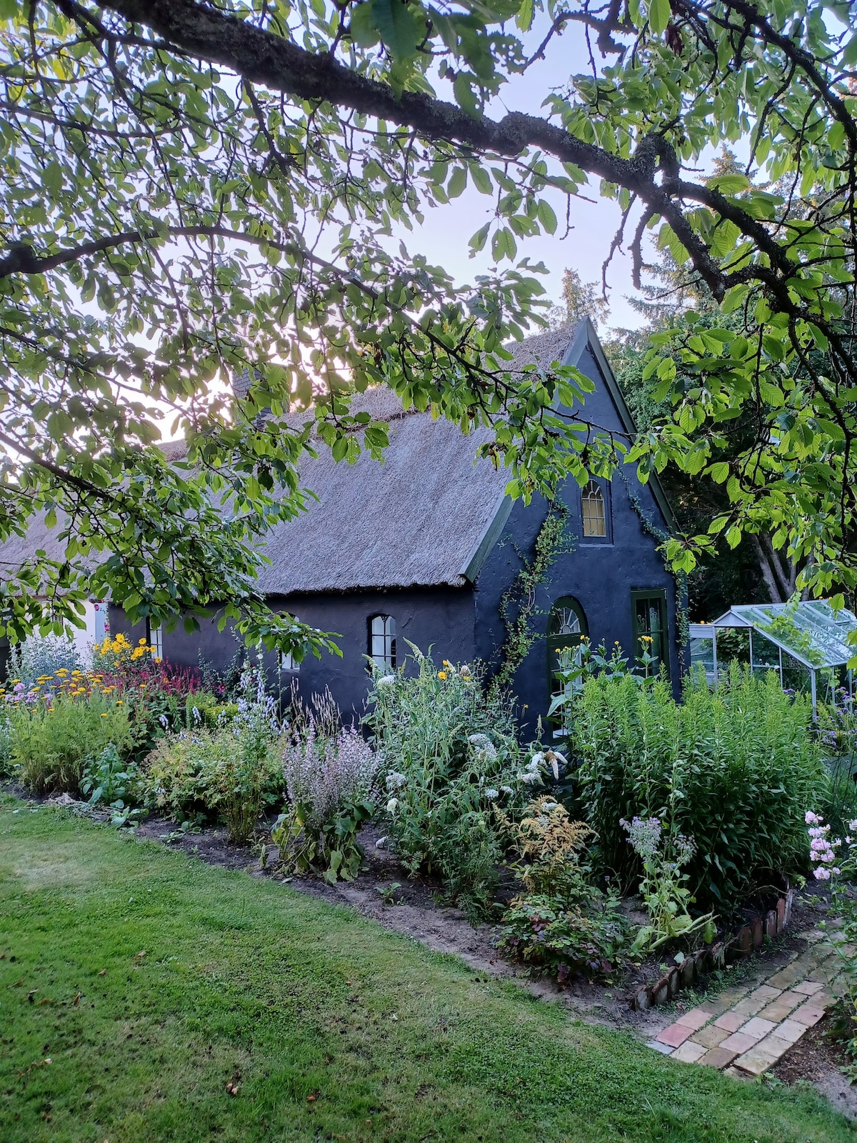 A charming black house with a thatched roof is surrounded by a vibrant flower garden. Lush greenery and colorful blooms create a natural border, while a pathway of bricks leads through the garden, emphasizing the peaceful, secluded setting.