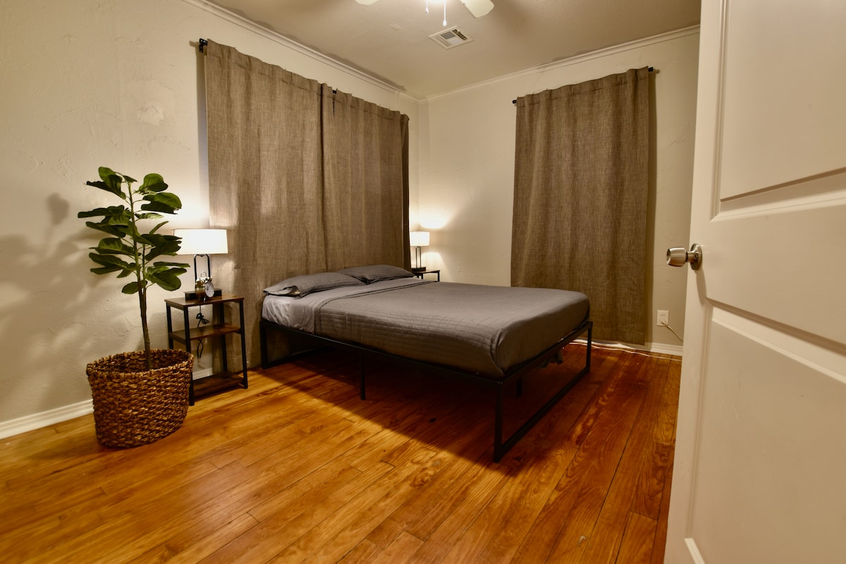 Un acogedor dormitorio con una cómoda cama tamaño queen y ricos pisos de madera. Disfruta de una amplia luz natural o corre las cortinas opacas para una noche tranquila. Una higuera de hoja de violín añade un toque de vegetación.