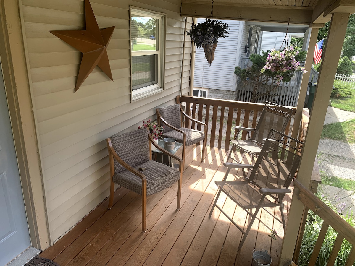 A front porch is shown with wooden flooring and beige siding. Two wicker chairs are positioned next to a table, with hanging plants and flower pots enhancing the space. A star decoration is mounted on the wall, and the view includes greenery and a nearby sidewalk.