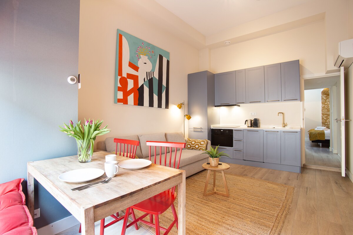 A modern dining area is displayed, featuring a wooden table set with two plates and a cup. Red chairs surround the table. The adjacent living space includes a cozy gray sofa and an artistic wall painting. A fully equipped kitchen is visible in the background.