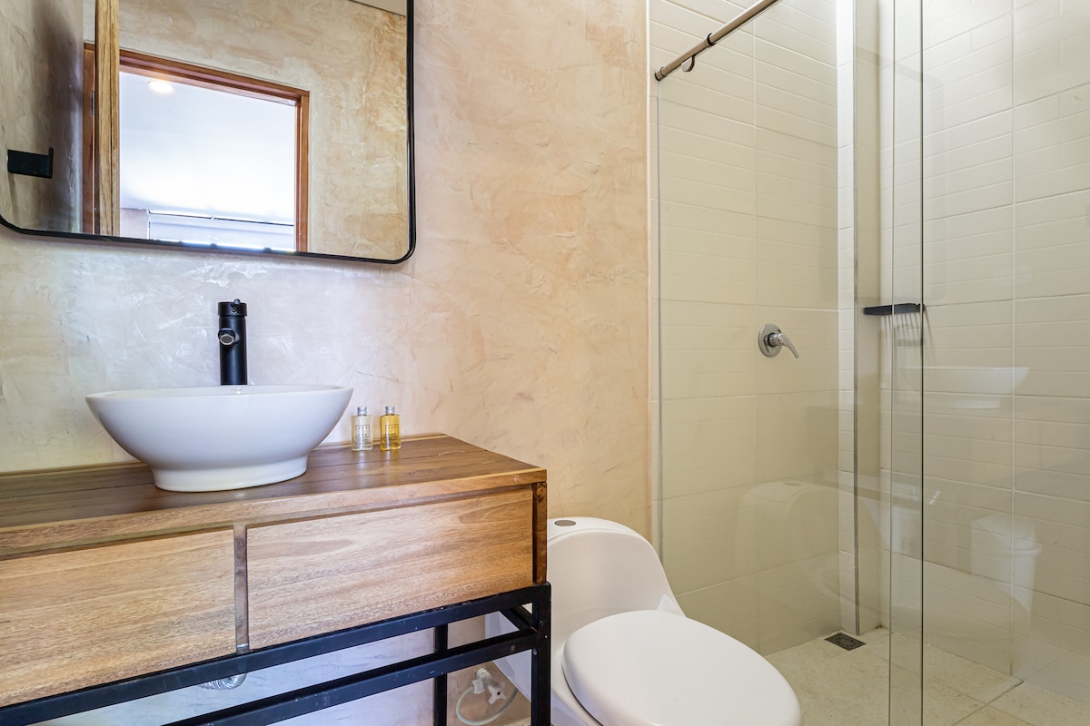A modern bathroom features a glass-enclosed shower with a rainfall showerhead and sleek tile walls. A large, circular basin rests atop a wooden vanity with an open frame, complemented by a minimalist mirror above. Natural light brightens the space through a nearby window.