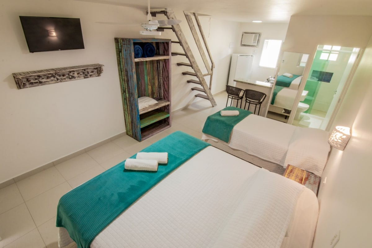 A well-lit room features two spacious beds adorned with turquoise and white linens. A colorful shelving unit is stocked with towels. A small dining table and chairs are positioned in the corner, with a television mounted on the wall. A bathroom is visible through a door.