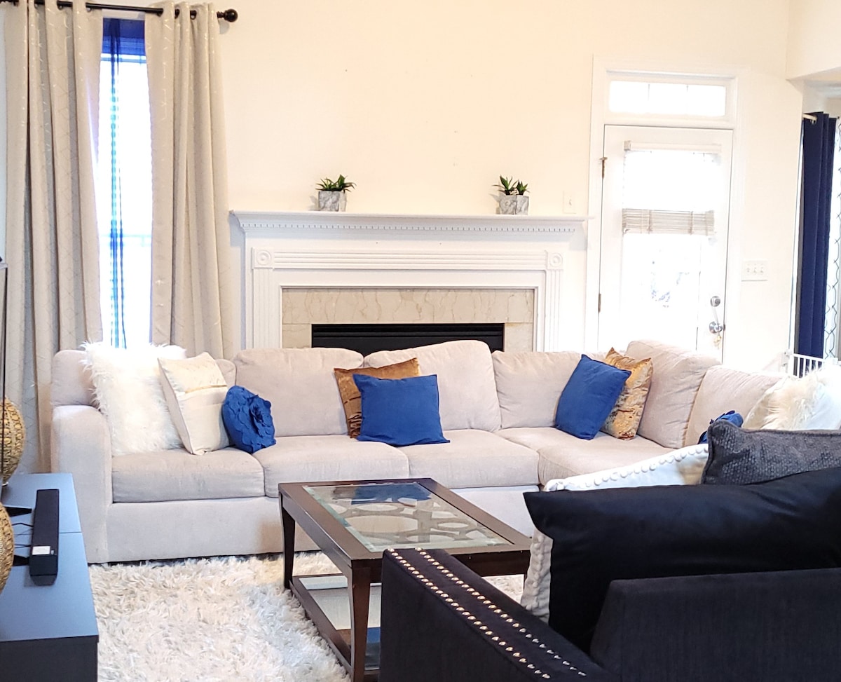 A spacious living area is furnished with a large, light-colored sectional sofa adorned with assorted blue and white pillows. A dark wood coffee table sits on a plush rug, and a decorative fireplace is framed by elegant curtains and potted plants.