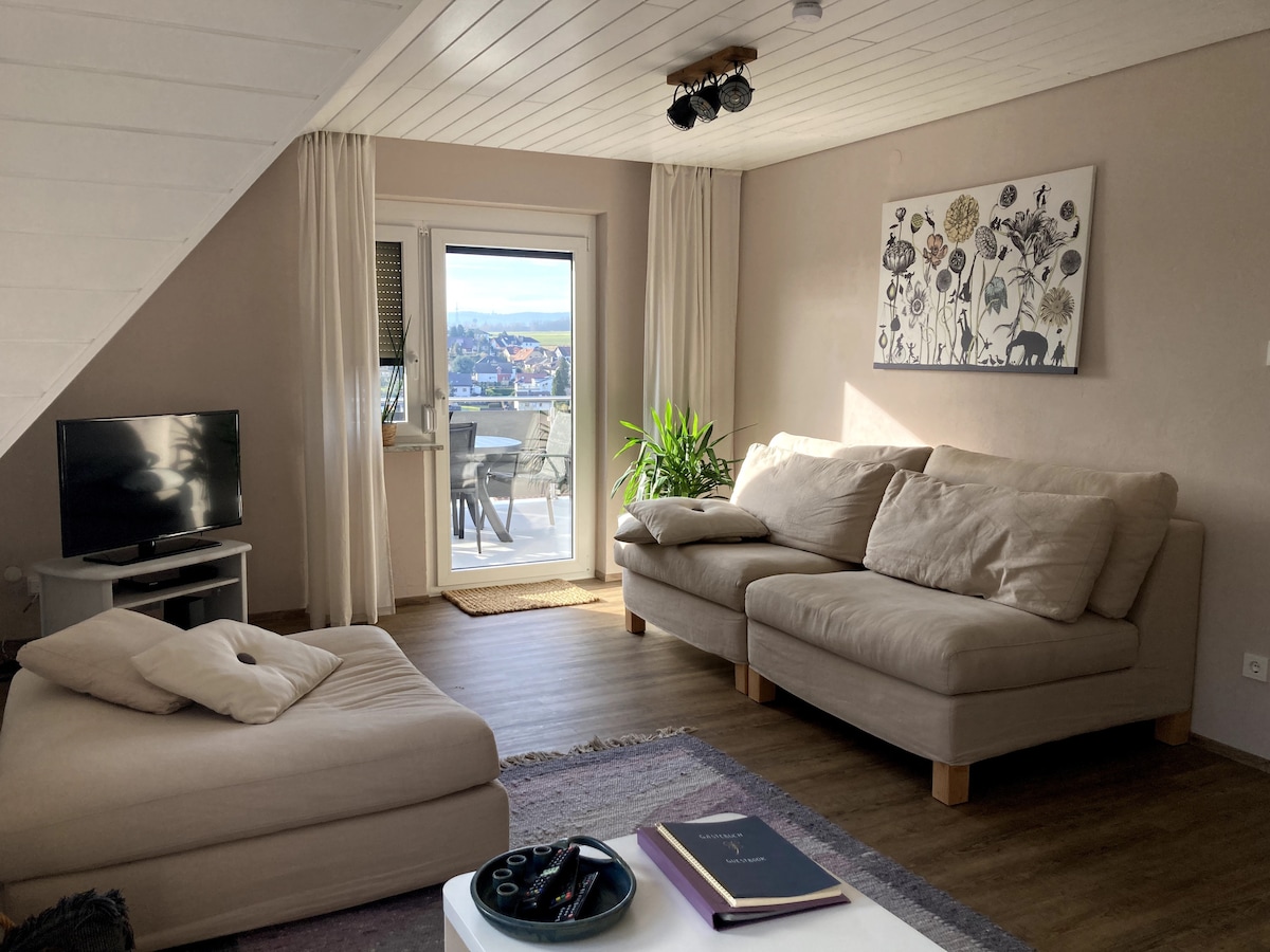 A living area features a large sectional sofa and a cozy armchair, complemented by a light wooden coffee table. Natural light floods in through a glass door leading to the balcony, where distant views can be enjoyed. A flat-screen TV is positioned against the wall.