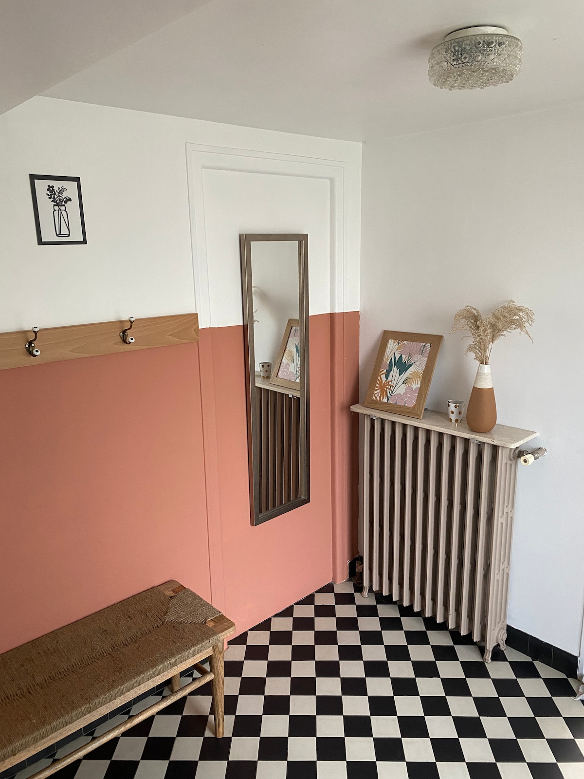 A welcoming entryway features a two-tone wall design, with peach and white segments. A mirror hangs above a wooden bench, complemented by coat hooks. Decorative elements include a potted plant and a vase with dried foliage, set against a contrasting black and white checkered floor.