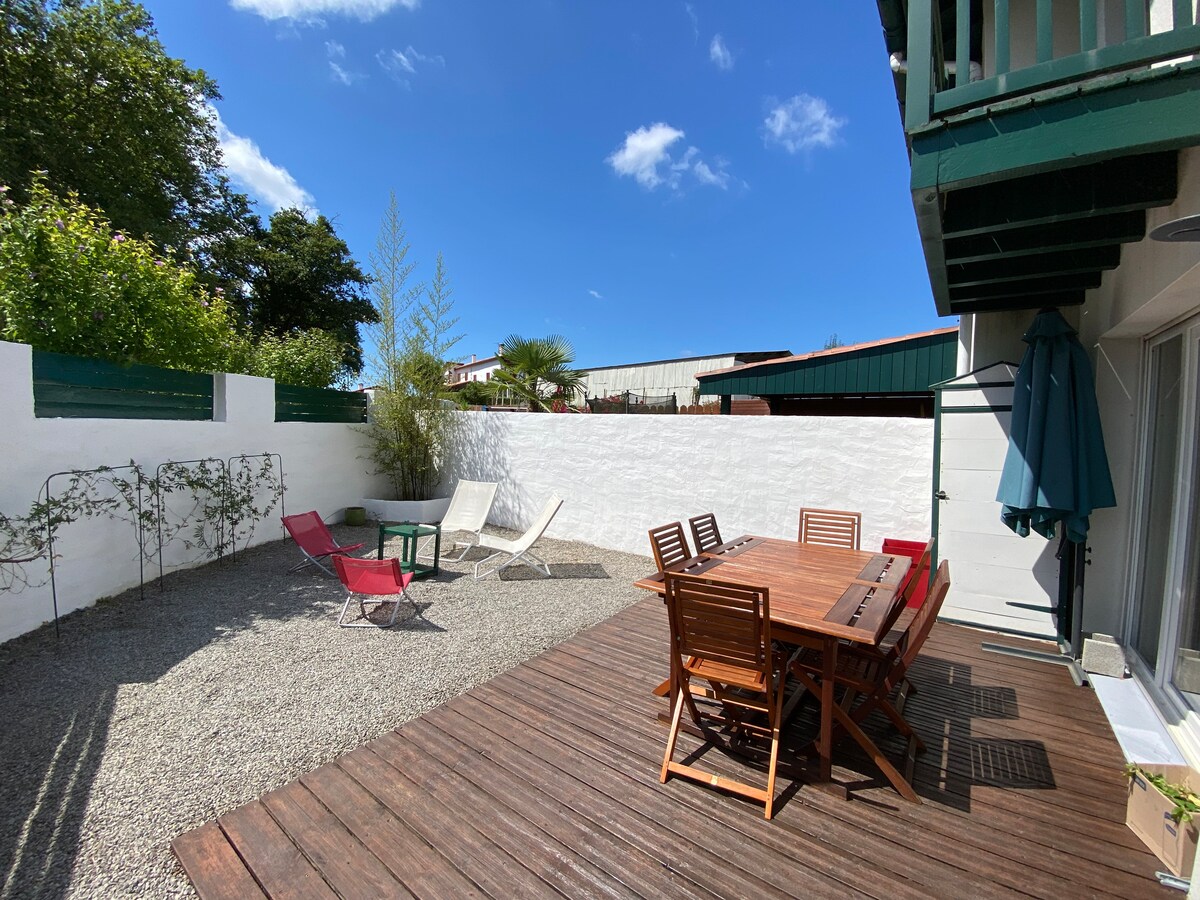 An outdoor space is showcased, featuring a wooden deck and a dining table surrounded by several chairs. Two red lounge chairs are positioned nearby, along with a small green table. A white wall encloses the area, enhanced by greenery and a blue sky overhead.