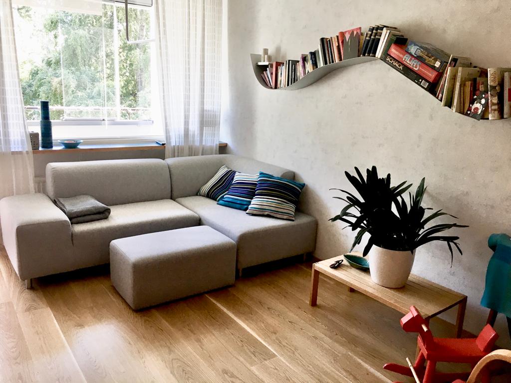 A modern living area features a light grey sectional sofa adorned with striped cushions. A wooden coffee table sits nearby, accompanied by a potted plant. Shelving along the wall displays books, while soft natural light filters through a large window, creating a bright and welcoming space.