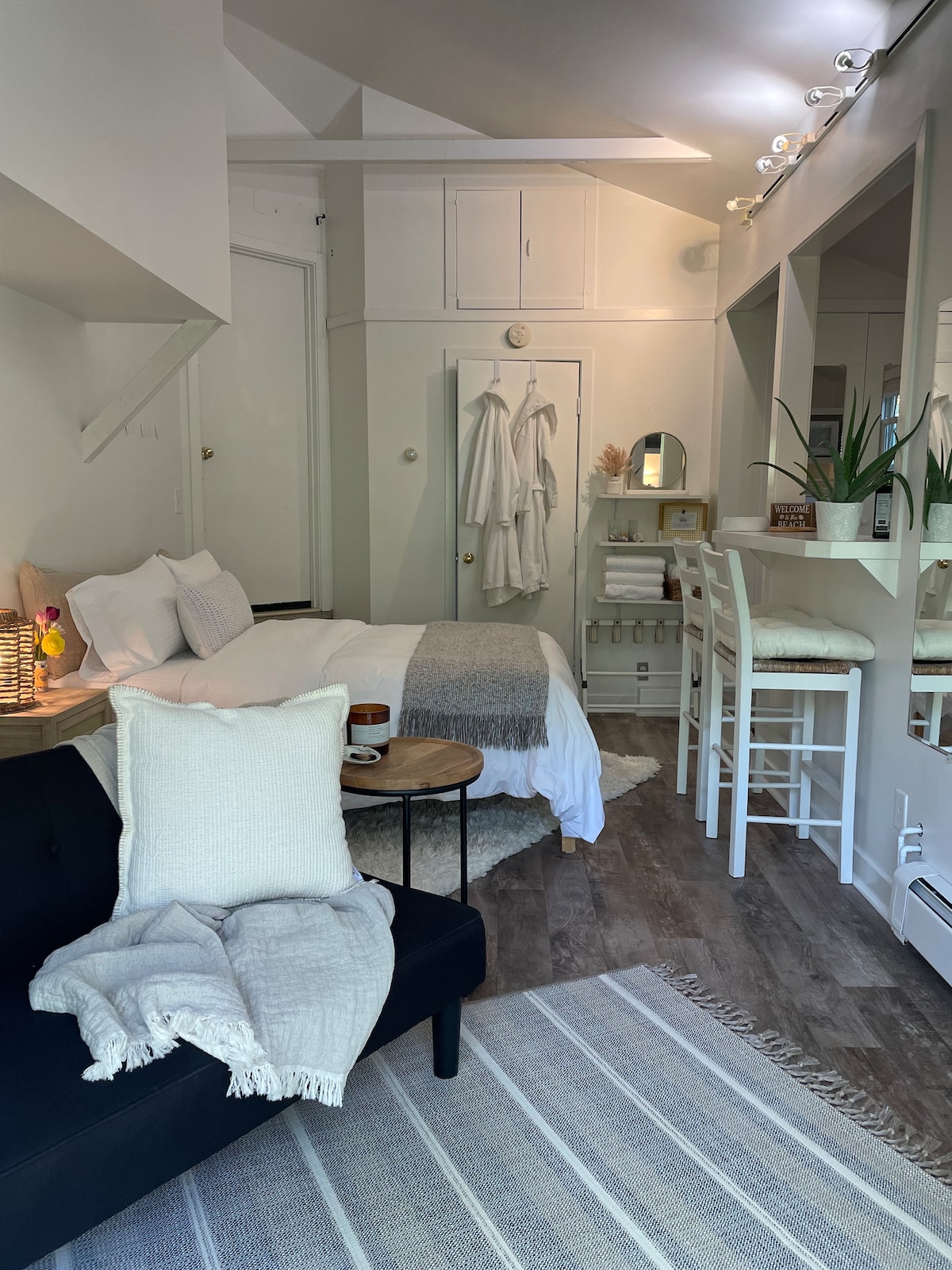 A cozy studio apartment features a spacious bed layered with a neutral quilt and decorative pillows. A small seating area with a black sofa, a side table, and a soft throw blanket invites relaxation. Natural light streams in, highlighting a kitchenette and seating bar in the background.