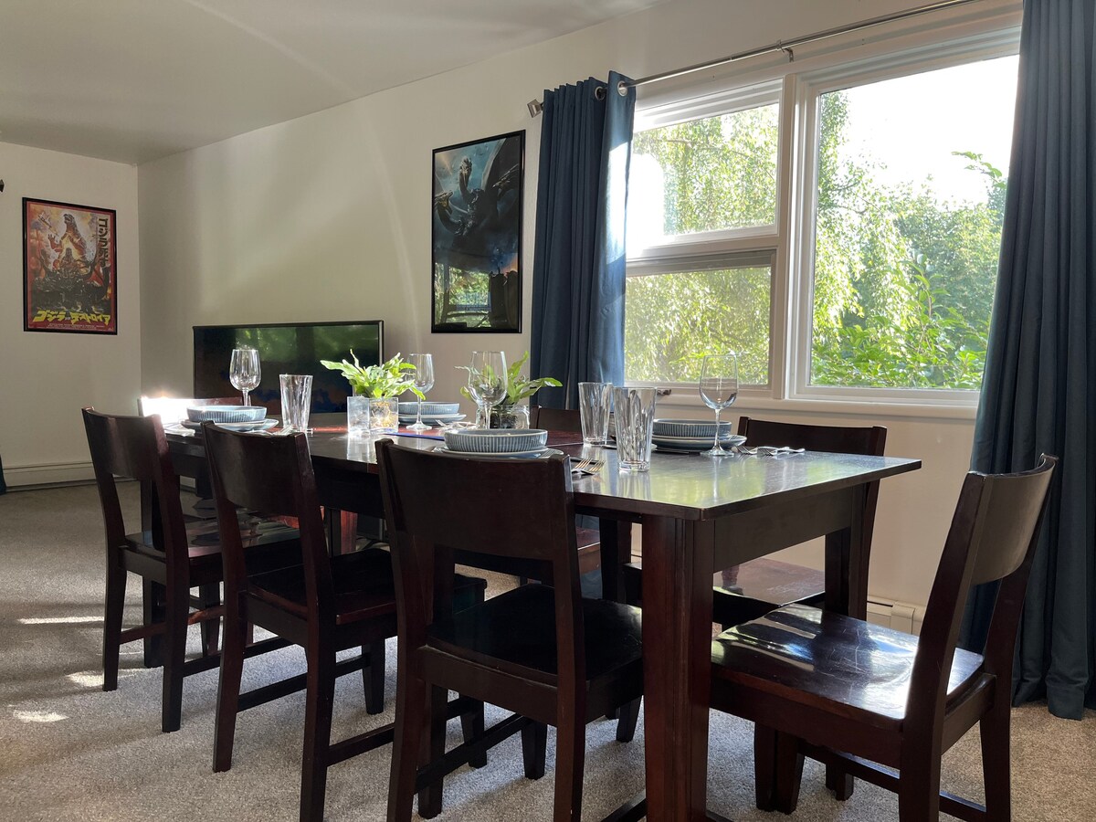 A spacious dining area features a large wooden table set for dining with plates, glasses, and cutlery. Natural light filters through a large window adorned with blue curtains, illuminating the surrounding chairs. Artwork is displayed on the walls, adding visual interest to the space.