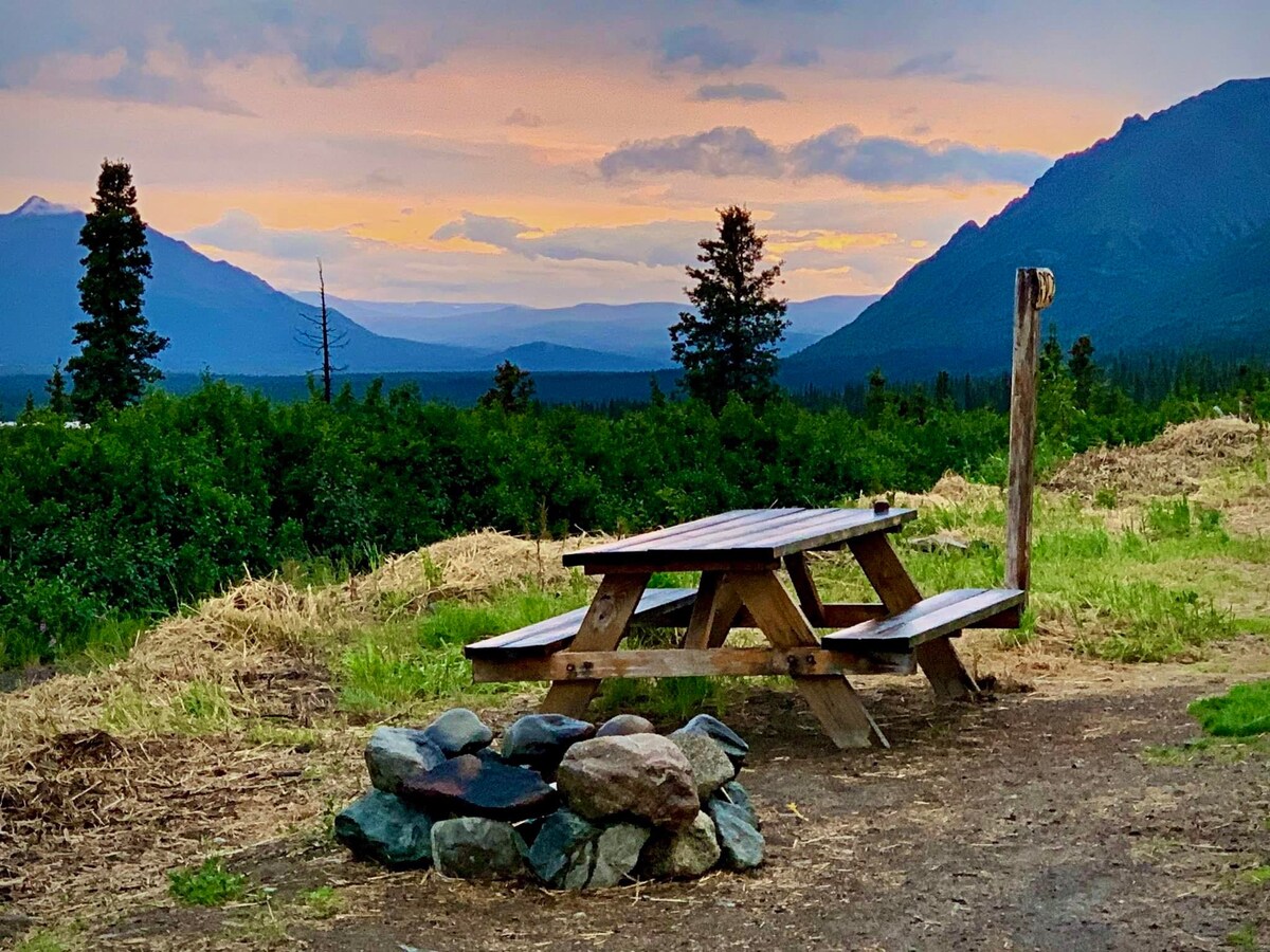 A rustic picnic table is positioned near a fire pit, surrounded by stones. The scene features expansive views of mountains and a colorful sunset sky, with lush greenery in the foreground. Soft clouds drift above, adding depth to the tranquil landscape.