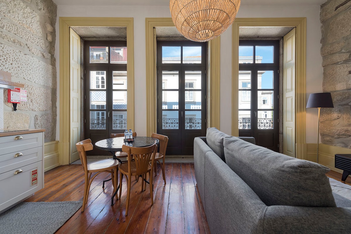 A cozy living area is revealed, featuring large windows that allow natural light to illuminate the space. A round dining table with two wooden chairs is positioned next to the window, while a gray sofa is situated nearby. The wooden flooring adds warmth to the environment.