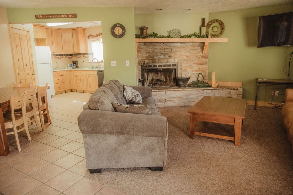 The living area features a cozy seating arrangement with a spacious sofa and a wooden coffee table. A stone fireplace adds warmth, while a television is mounted on the wall. The open kitchen area is visible, showcasing wooden cabinetry and appliances.