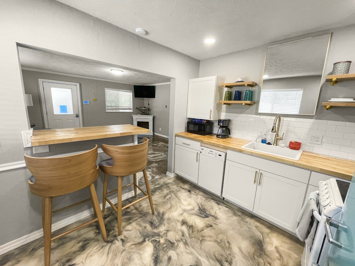A modern kitchen is showcased, featuring light wood countertops and white cabinetry. Two high-backed bar stools are positioned at a small counter. A mirror reflects the natural light from a nearby window, and appliances are neatly arranged along the wall.