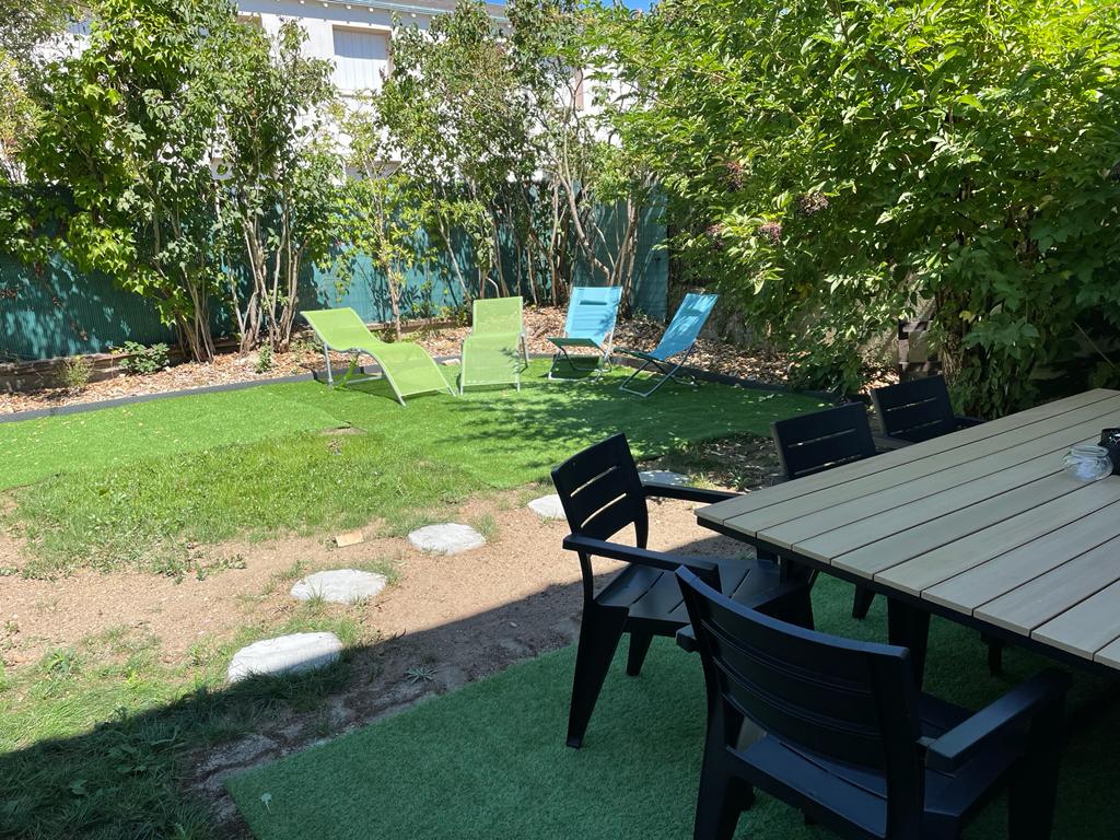 A garden area features manicured grass and a variety of seating options, including a wooden dining table with several chairs. Four lounge chairs in soft hues are positioned on the lawn, surrounded by lush greenery offering privacy and shade.