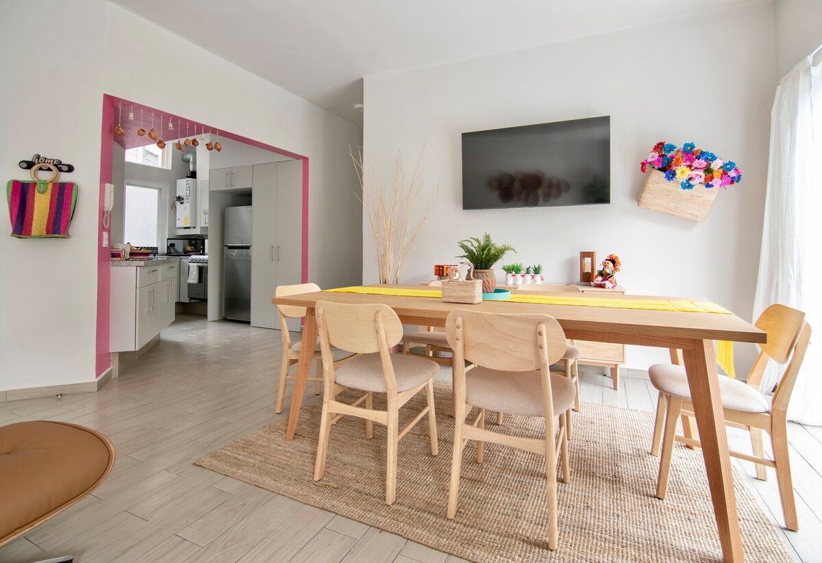 A modern dining area features a wooden table surrounded by light-toned chairs. A large flat-screen TV is mounted on the wall. Natural light enters through floor-to-ceiling windows, illuminating a woven rug beneath the table. The kitchen is visible in the background.