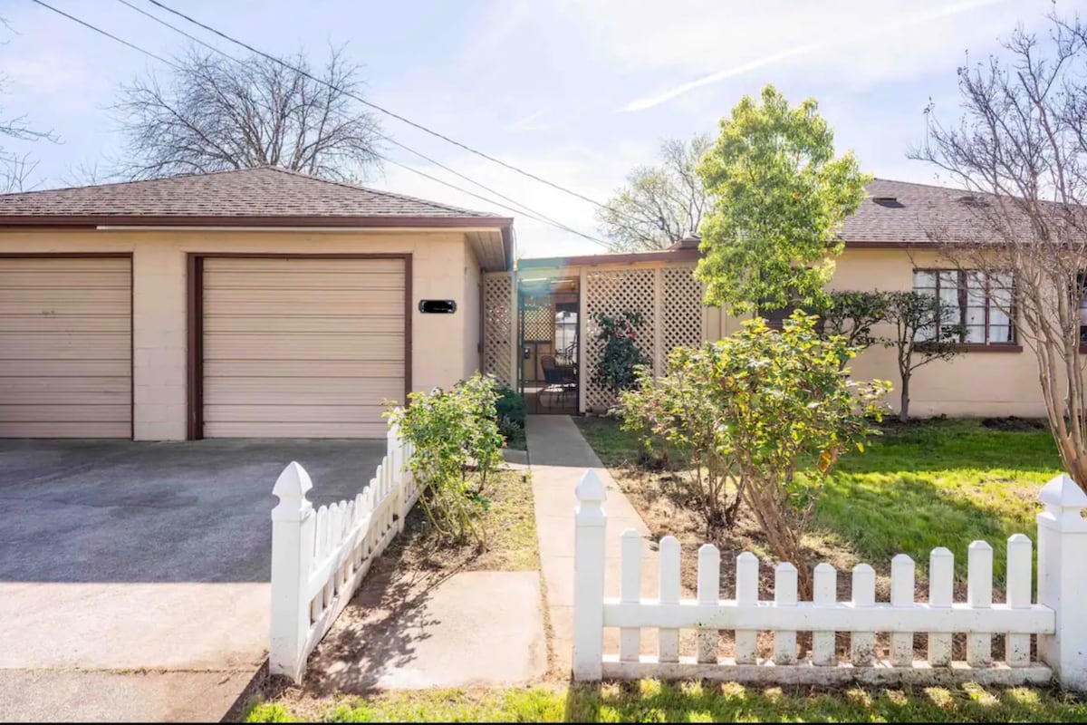 The exterior of the home features a beige block façade with a brown shingle roof. A small white picket fence borders the front yard, which contains low shrubbery and a pathway leading to the entrance. Two garages are visible on the left.
