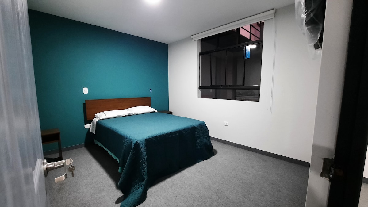 A simple sleeping area features a spacious bed dressed in a teal blanket. A large window allows natural light to fill the room, while the walls are painted in a calming teal. A bedside table is positioned next to the bed.