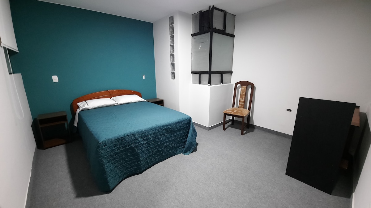The sleeping area features a spacious bed with a teal coverlet, complemented by a wooden headboard. A single wooden chair and a dark cabinet provide functional furnishings. Natural light filters through a window, illuminating the room's neutral tones.