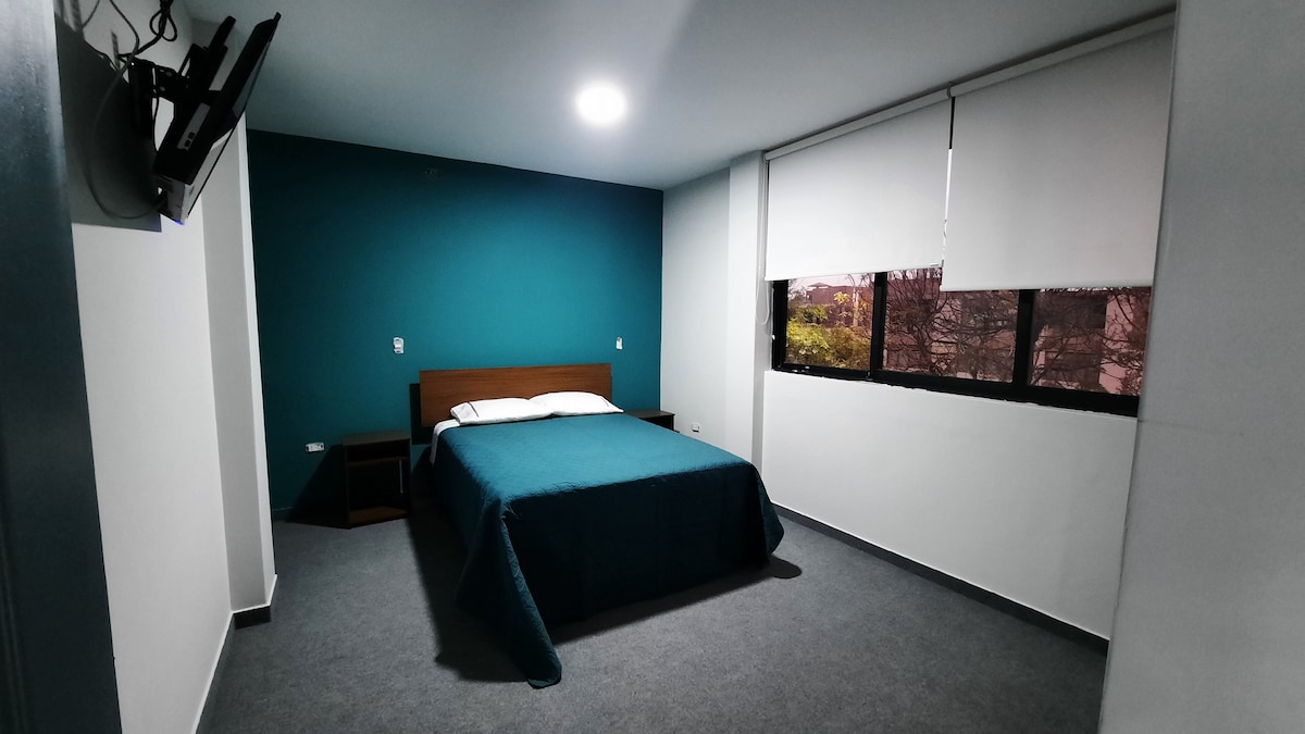 A spacious bedroom features a large bed topped with a teal blanket. The walls are painted in a deep teal color, and a flat-screen television is mounted on the wall. Natural light filters through a large window adorned with white blinds.