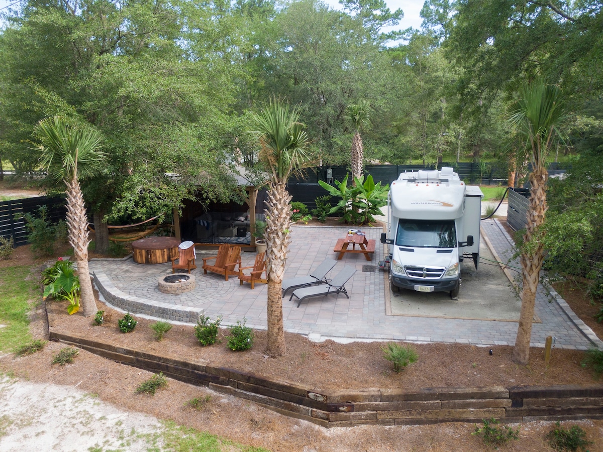 An aerial view captures the RV site featuring a spacious, paved patio area surrounded by palm trees. The site includes a fire pit, Adirondack chairs, and a small hot tub, all set against a backdrop of lush greenery. An RV is parked nearby.