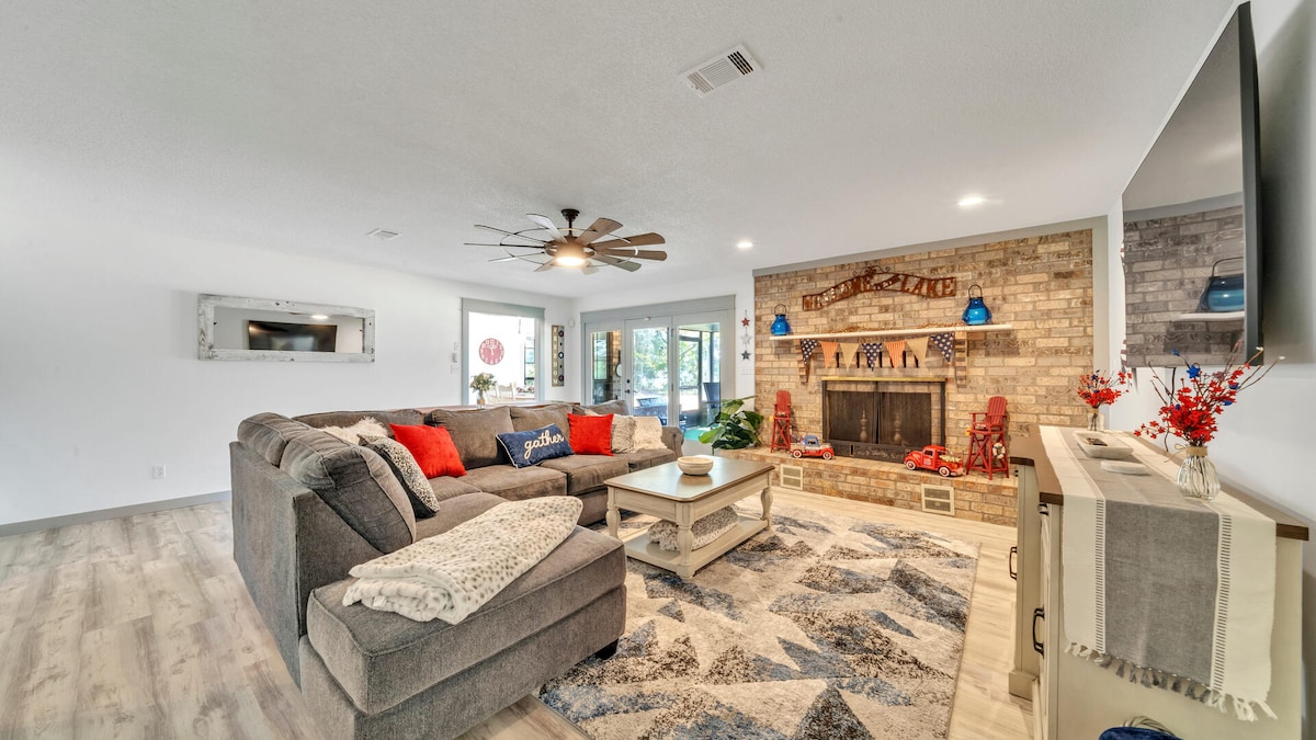 A spacious living room features a large sectional sofa adorned with red and blue cushions. A coffee table sits in front of the cozy stone fireplace, while a television is mounted on the wall. Natural light streams in through nearby sliding doors, enhancing the inviting ambiance.