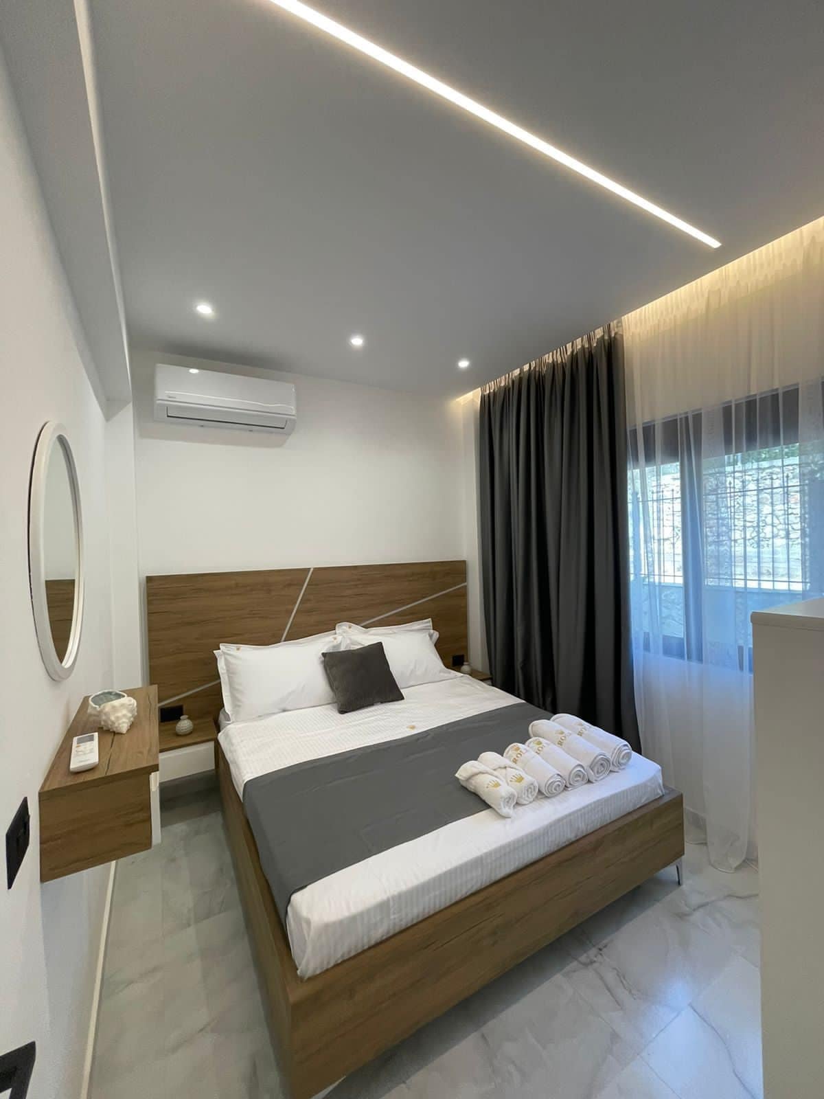 A cozy bedroom features a spacious bed adorned with neatly arranged towels. Large windows allow natural light to fill the room, with sheer curtains softening the light. Warm wooden accents complement the minimalistic design, and an air conditioning unit is mounted on the wall.