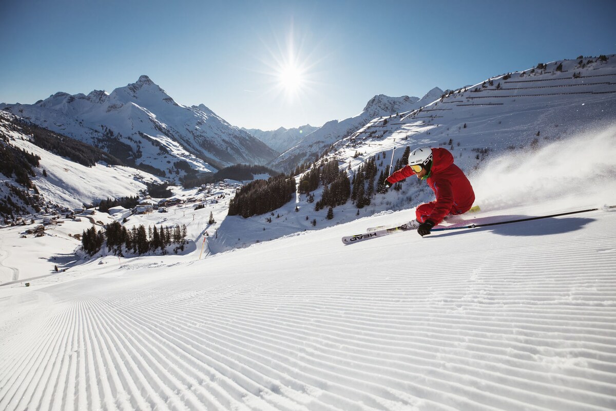 A skier in a vibrant red suit gracefully navigates down a groomed slope, surrounded by pristine snow-covered mountains. The bright sun illuminates the scene, casting a cheerful glow over the landscape, which features valleys and vast stretches of untouched snow.