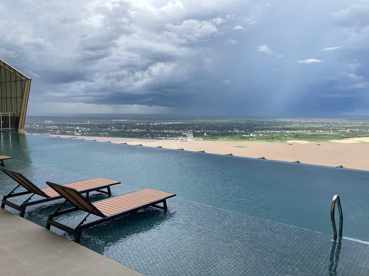 An infinity pool extends toward the horizon, reflecting the sky's dynamic cloud formations. Two wooden loungers are positioned nearby on the pool deck, offering a serene spot to relax and enjoy expansive views of the surrounding landscape.