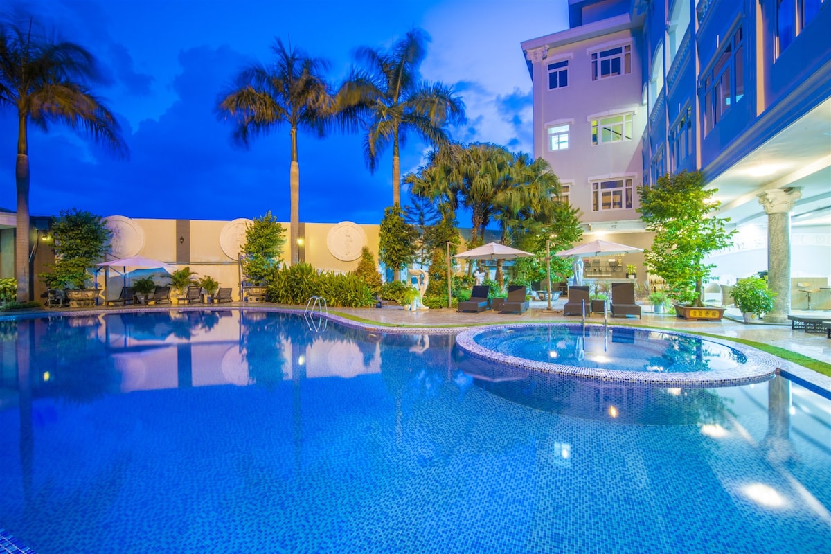 An outdoor swimming pool is framed by tropical palm trees, reflecting a tranquil blue hue under the evening sky. Sun loungers are arranged around the pool, accompanied by umbrellas, creating a relaxing atmosphere. Lush greenery adds to the serene ambiance of the hotel grounds.