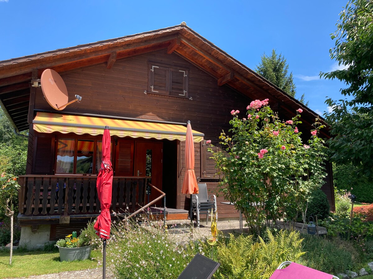 A charming wooden house with a sloped roof is surrounded by lush greenery and colorful blooming flowers. A covered porch features striped awnings, offering shaded seating. In the foreground, umbrellas provide shade for outdoor relaxation as the sun shines against a clear blue sky.